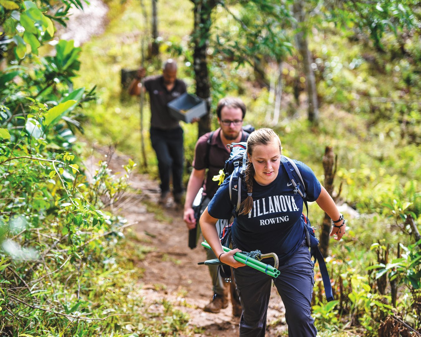 Female student in a Villanova T-shirt carrying water testing equipment and male student walk on a dirt path in Madagascar 