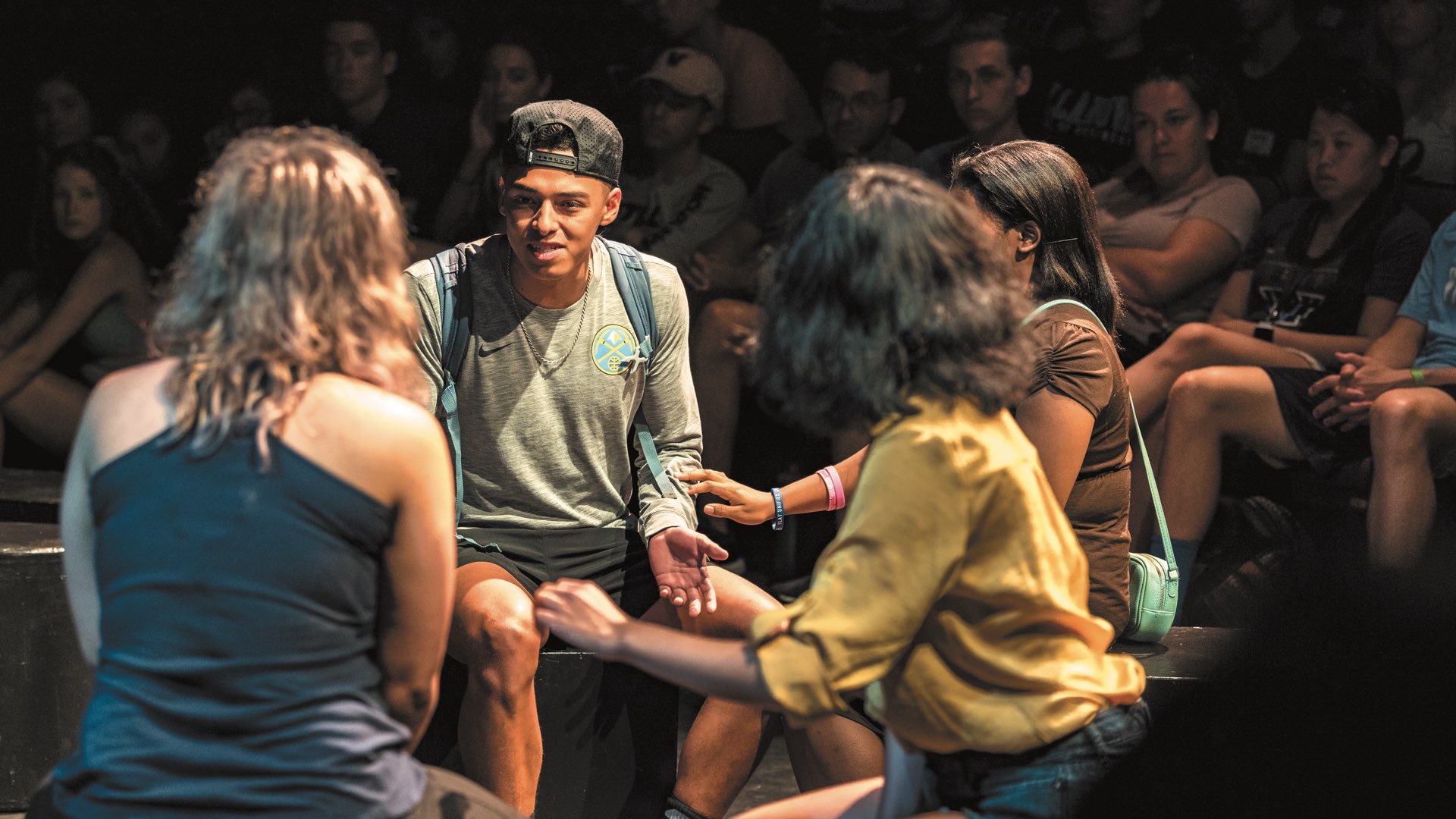 Backs of two female students talking with a male student wearing a gold chain and backwards baseball cap in Vasey Theatre