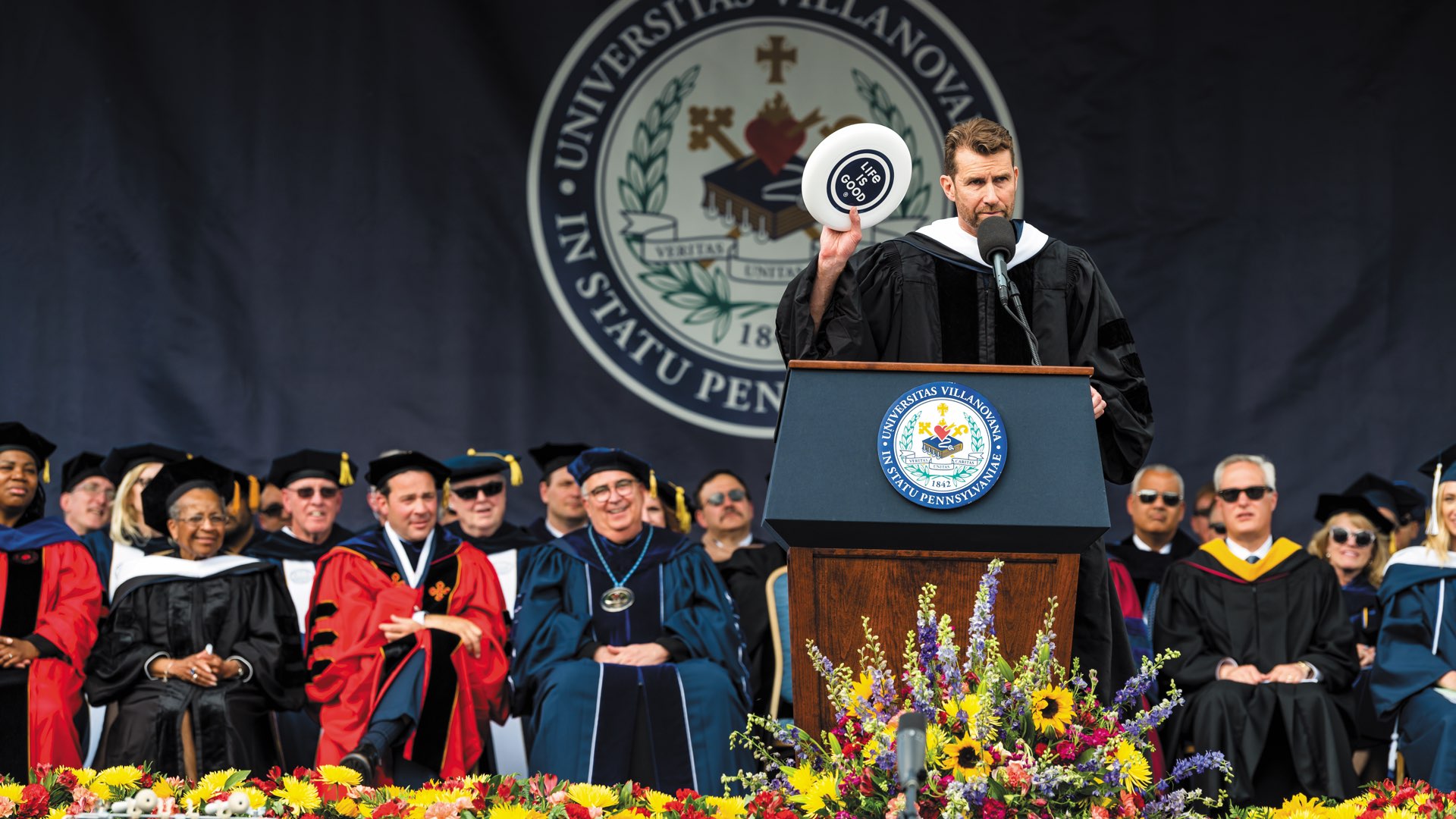 On stage, Commencement speaker Bert Jacobs at the podium in graduation regalia holding up a Life Is Good frisbee
