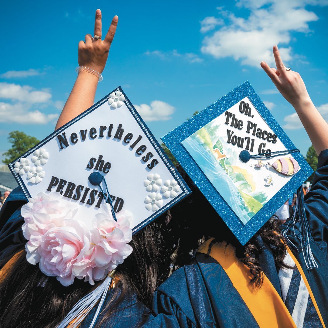 Two female students giving the peace sign, wearing caps that read “Nevertheless she persisted” and “Oh the places you’ll go”