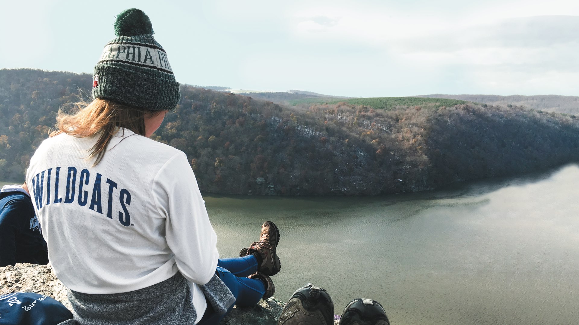 A female student in an Eagles wool cap and Wildcats shirt sits atop a mountain in hiking boots overlooking a scenic view